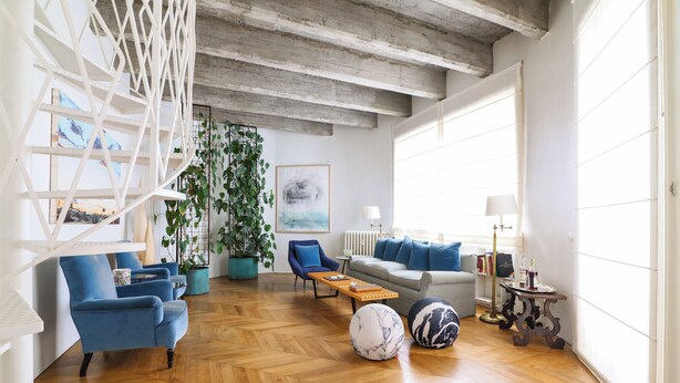 A spacious living room with a herringbone wooden floor, a blue sofa set, a coffee table, and a large potted plant.