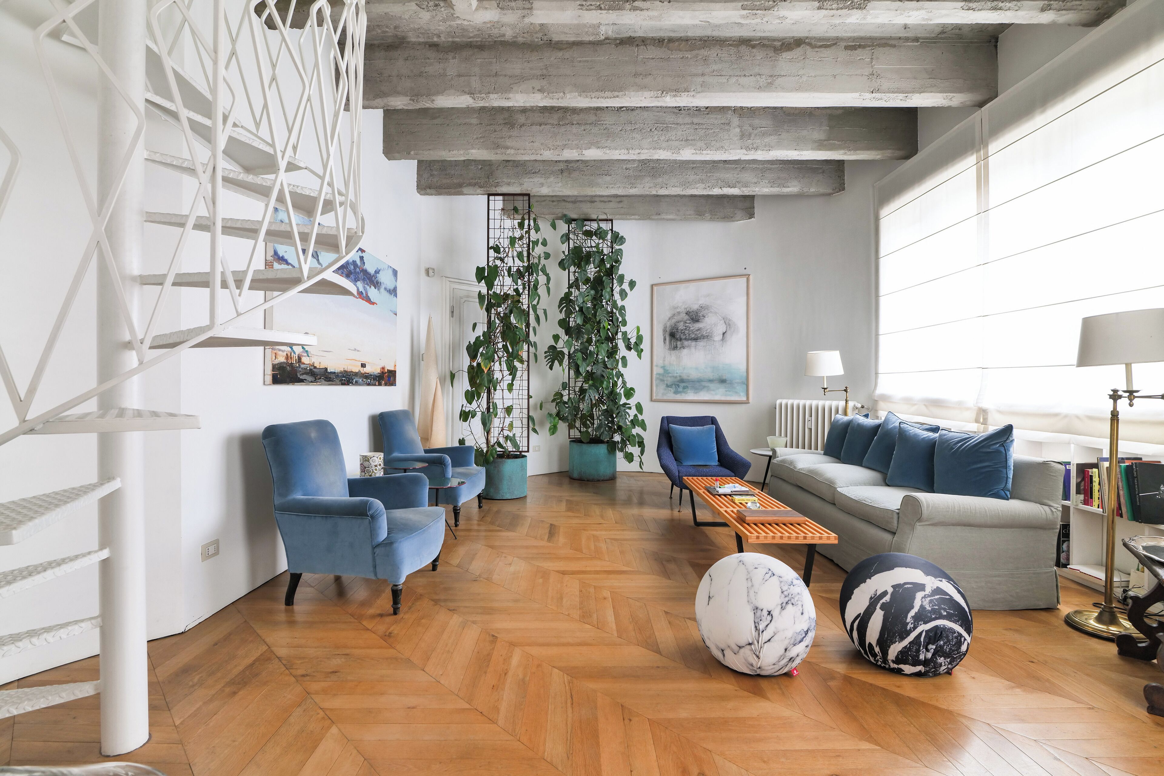 A modern living room with a herringbone wooden floor, blue armchairs, a grey sofa, and a coffee table. There are potted plants, a large abstract painting, and a white staircase with a geometric design.