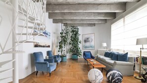 A modern living room with a herringbone wooden floor, blue armchairs, a grey sofa, and a coffee table. There are potted plants, a large abstract painting, and a white staircase with a geometric design.