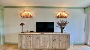 A wooden entertainment center with a flat-screen TV, two wall-mounted lamps, a vase with dried flowers, and a remote control on a light-colored wooden floor. - Cozy 2 bedroom flat with beautiful views (Cortina d'Ampezzo)