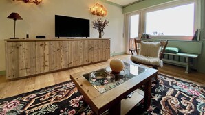 A living room with a wooden cabinet, a flat-screen TV, a glass coffee table with a decorative item, a patterned rug, and a chair with a fluffy cushion. - Cozy 2 bedroom flat with beautiful views (Cortina d'Ampezzo)