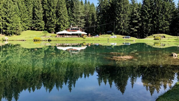 A lodge with a large building surrounded by dense forest, a calm lake reflecting the trees, and a clear blue sky. - Cozy 2 bedroom flat with beautiful views (Cortina d'Ampezzo)