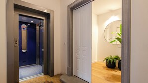 Een lift met een digitaal display, een witte deur met een spiegel, en een houten vloer met een vloerkleed en potplanten.
