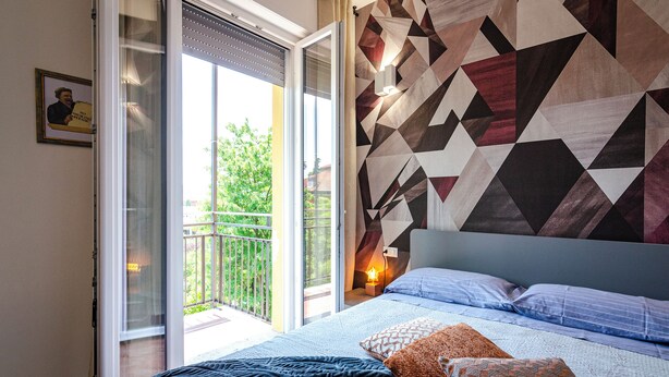 Ein Schlafzimmer mit Bett, Kissen, einer Wand mit geometrischem Muster, einem Nachttisch mit Lampe und einer Glastür zum Balkon.