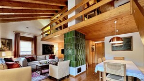 A spacious living room with a high wooden ceiling, a green glass fireplace, a dining area with a table and chairs, and a sofa set. - Classy 3 bedroom few steps from centre (Cortina d'Ampezzo)