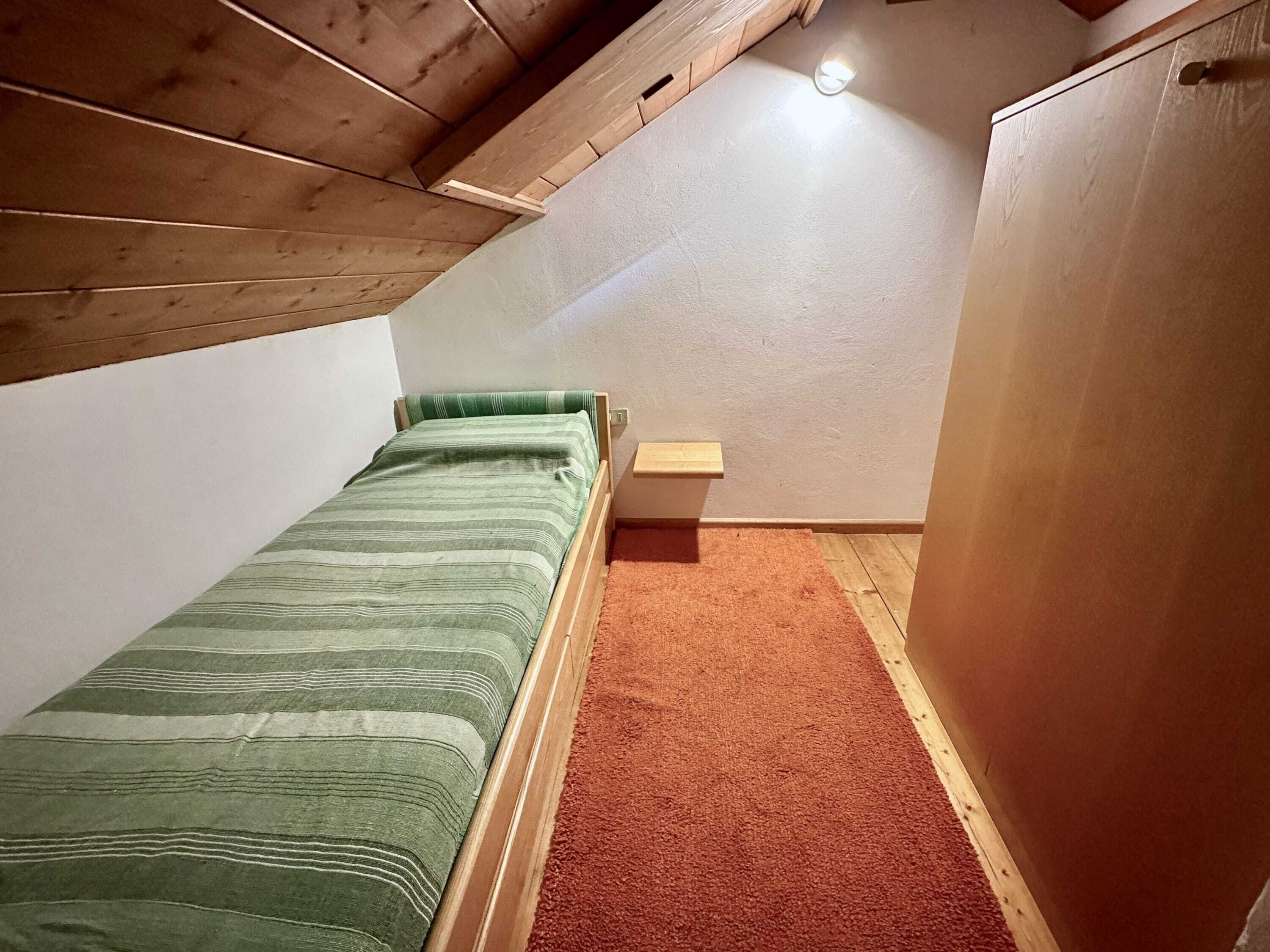 A small, single-bed room with a sloped ceiling, a wooden headboard, a green-striped bedspread, a small bedside table, and a red carpet.