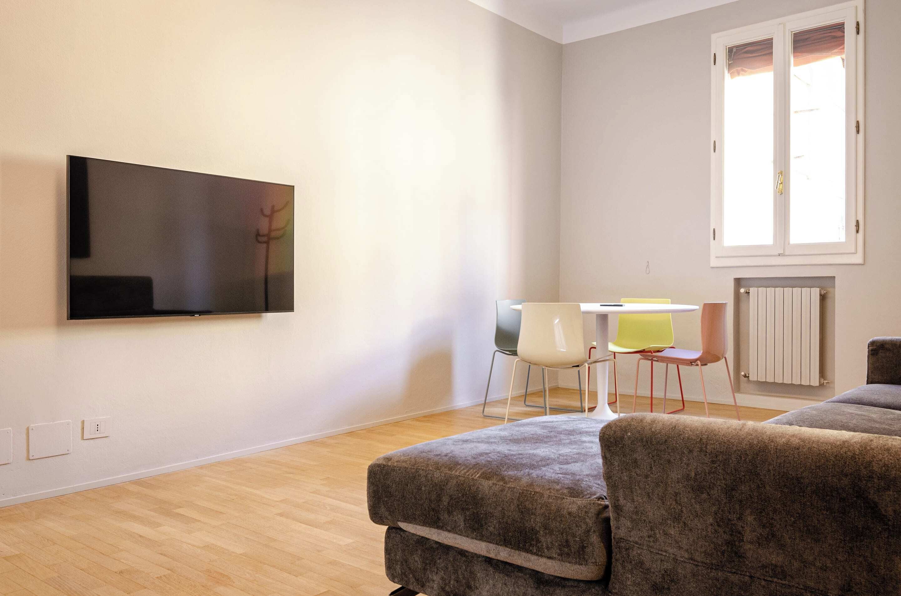 A modern living room with a flat-screen TV mounted on the wall, a small round table with two chairs, and a radiator on the wall.