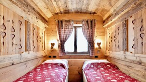 Ein Holzhütten-Zimmer mit zwei Betten, Karo-Vorhängen, herzförmigen Ausschnitten an den Wänden und einem Fenster mit Sonnenschein.