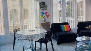 Una habitación con una mesa redonda, dos sillas, un sofá con un cojín de arcoíris y una pequeña mesita de noche con libros.