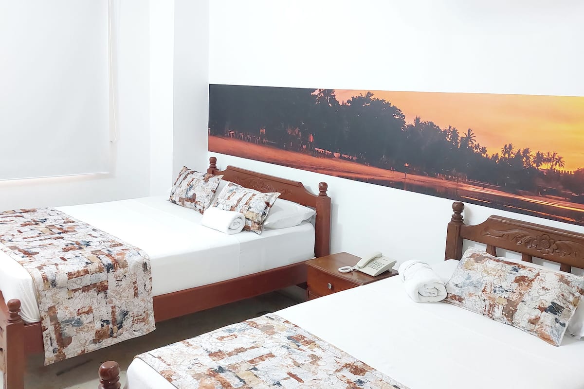 Une chambre d’hôtel avec deux lits, chacun doté d’une tête de lit en bois et d’une couvre-lit à motifs. Un grand tableau représentant un coucher de soleil est accroché au mur, derrière les lits.