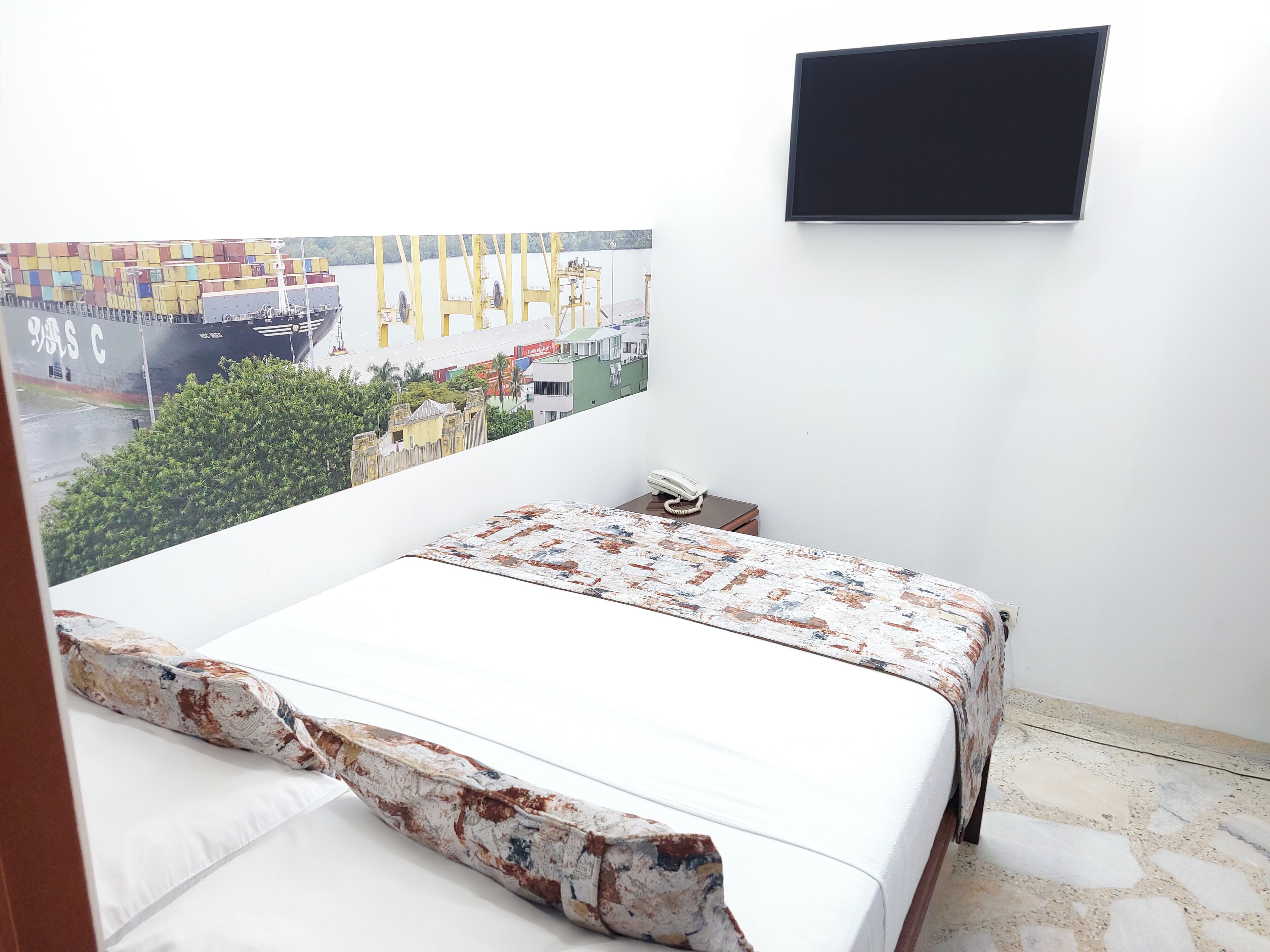 Un dormitorio con una cama, teléfono, televisor y un amplio ventanal con vista a un barco y edificios.