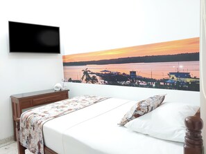 Une chambre d’hôtel comprenant un lit, une table de chevet, un téléviseur à écran plat fixé au mur et un grand tableau représentant un coucher de soleil sur une scène côtière.