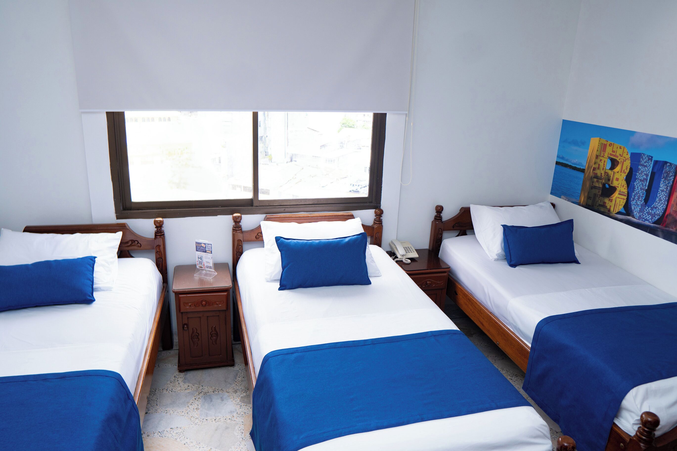 A hotel room with three single beds, each with blue bedding and white pillows. There is a wooden headboard, a bedside table with a phone, and a window with blinds.