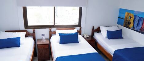 A hotel room with three single beds, each with blue bedding and white pillows. There is a wooden headboard, a bedside table with a phone, and a window with blinds.