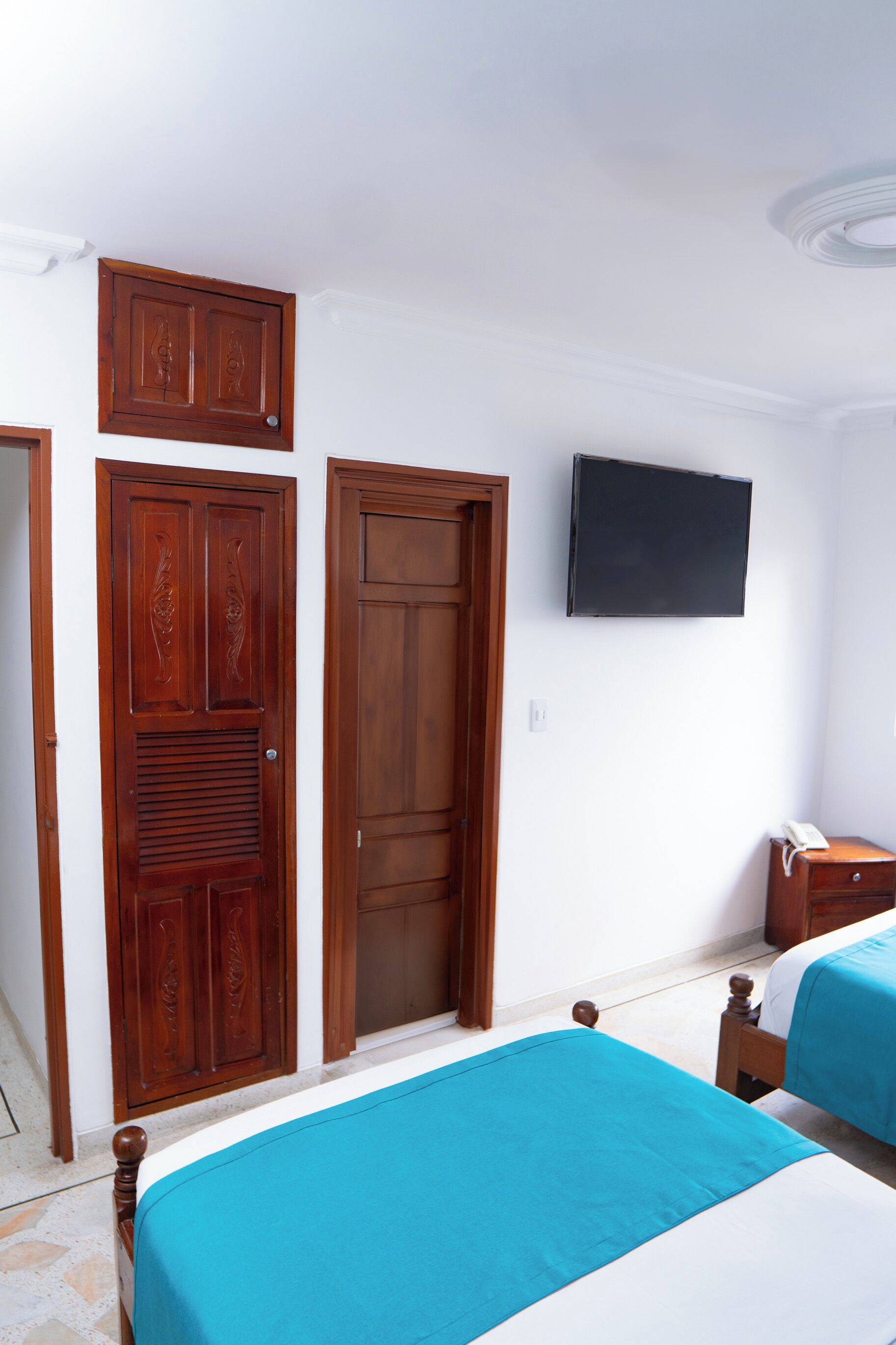 A bedroom with two beds, a wooden wardrobe, a television on the wall, and a bedside table.