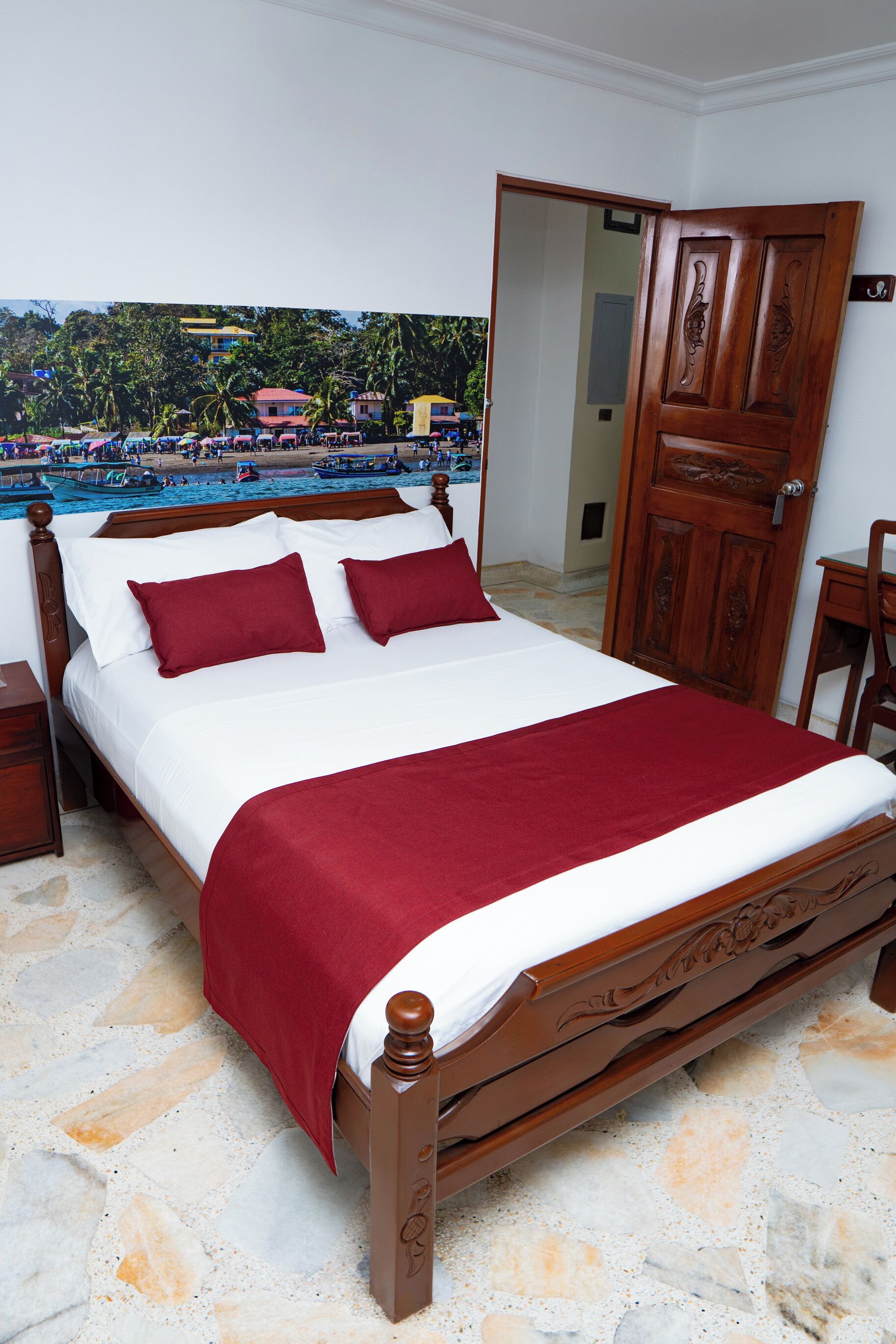 Una cama bien hecha con una colcha y almohadas rojas, un armario de madera y vistas a una playa y palmeras a través de una puerta abierta.