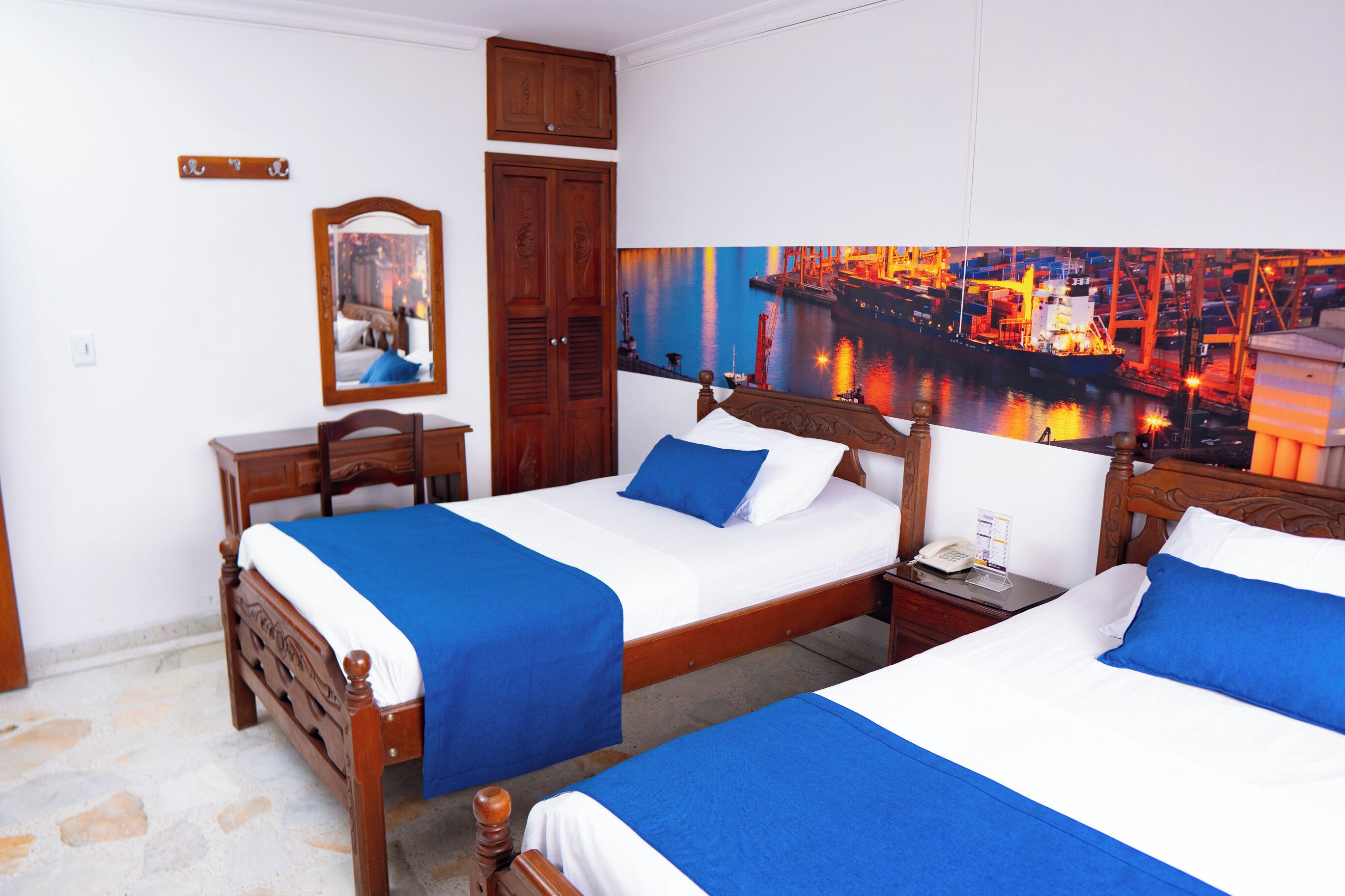 A hotel room with two wooden beds, each with a blue bedspread and white linens. There is a wooden nightstand with a telephone and a small table with a lamp. A large painting of a harbor scene is on the wall.