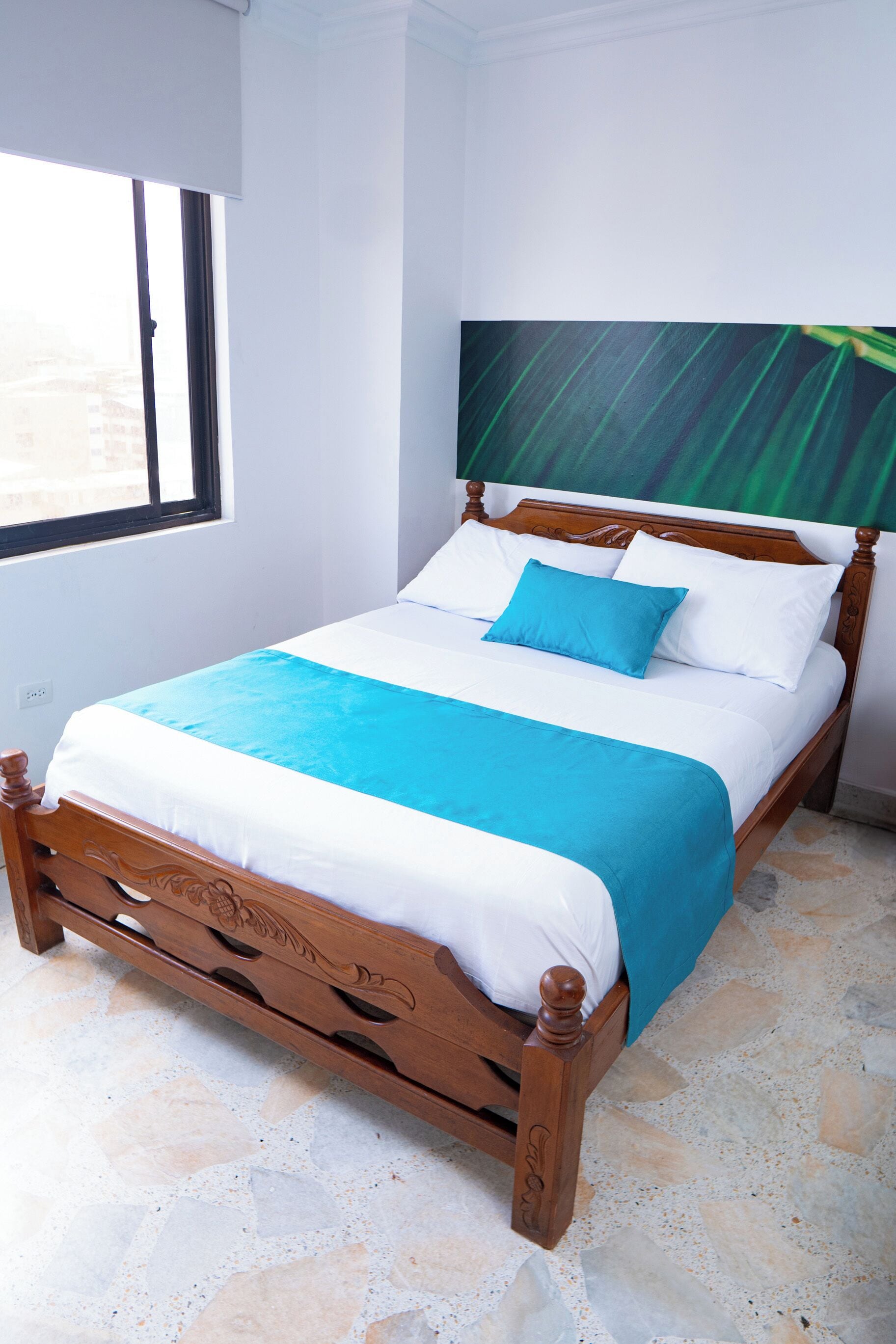 Una cama bien hecha con estructura de madera, colcha azul y blanca y una sola almohada azul.