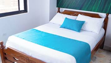 Una cama bien hecha con estructura de madera, colcha azul y blanca y una sola almohada azul.