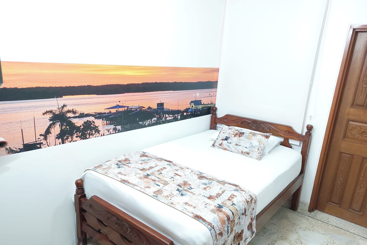 Une chambre à coucher avec un lit, une vue sur un coucher de soleil se reflétant sur une étendue d’eau, et une porte en bois.