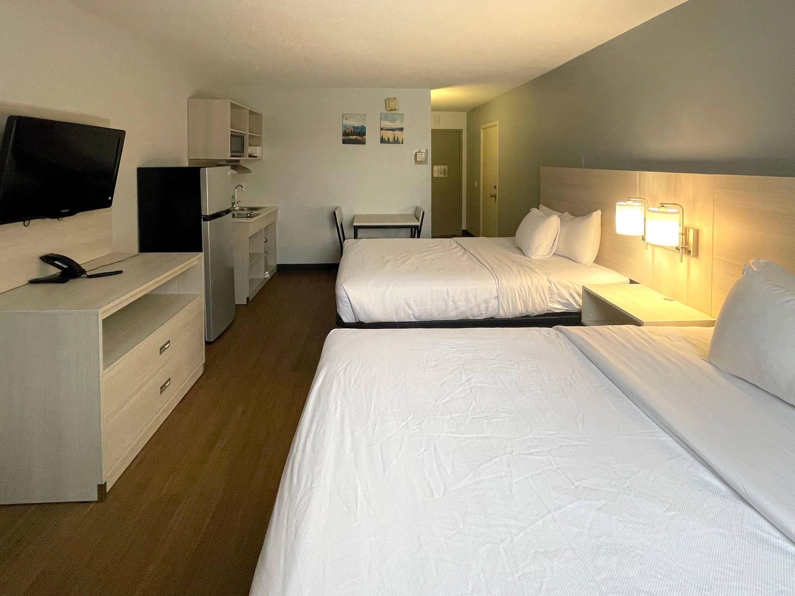 A hotel room with two beds, a desk with a chair, a television, a small kitchenette, and a bathroom door.