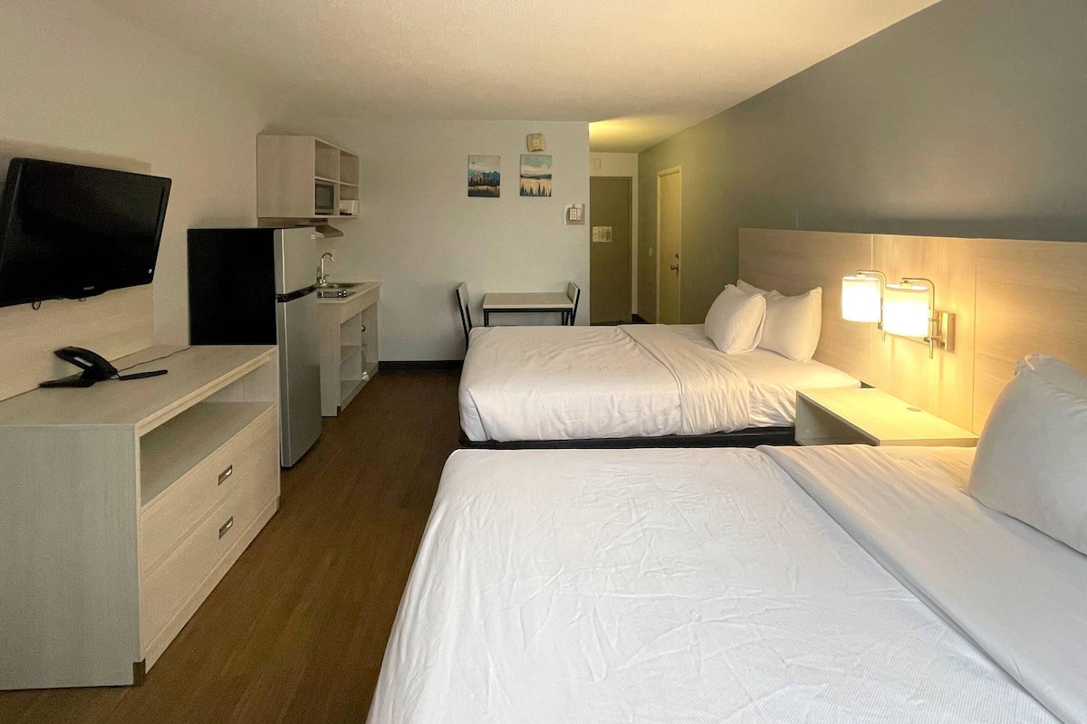A hotel room with two beds, a desk, a television, and a small kitchenette.
