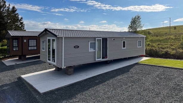 Il s’agit d’une petite maison mobile d’un seul étage, à l’extérieur gris et au toit recouvert de tuiles. Elle possède une porte d’entrée, une petite fenêtre et une porte donnant sur un porche. La maison mobile est garée sur une allée en gravier, avec une zone herbeuse en arrière-plan.