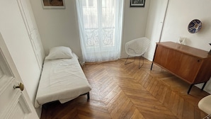 A small, well-lit room with a herringbone wooden floor, a single bed, a chair, a small desk, and a window with sheer curtains. - Apartment in lively neighborhood near Marais.  (Paris)