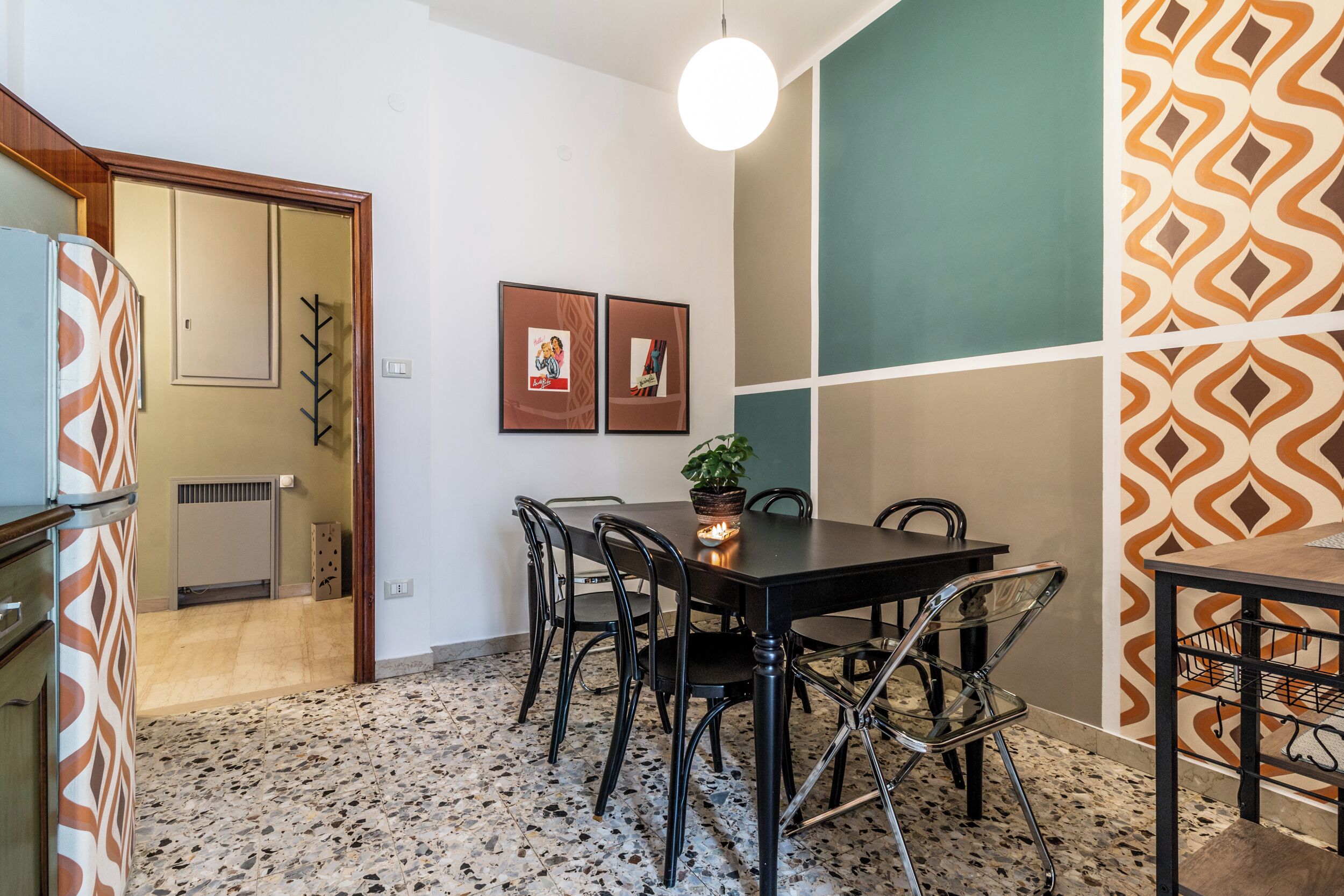 Una zona de menjador amb una taula negra, cadires metàl·liques i una planta en test. Hi ha quadres a la paret i una paret amb dibuixos decoratius a la dreta.