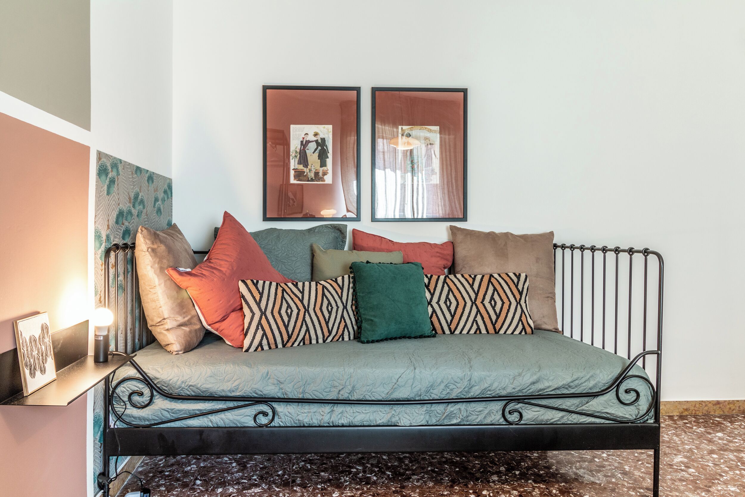Un lit avec un cadre en métal, agrémenté de coussins à motifs, un cadre photo fixé au mur et une petite table de chevet avec une lampe.