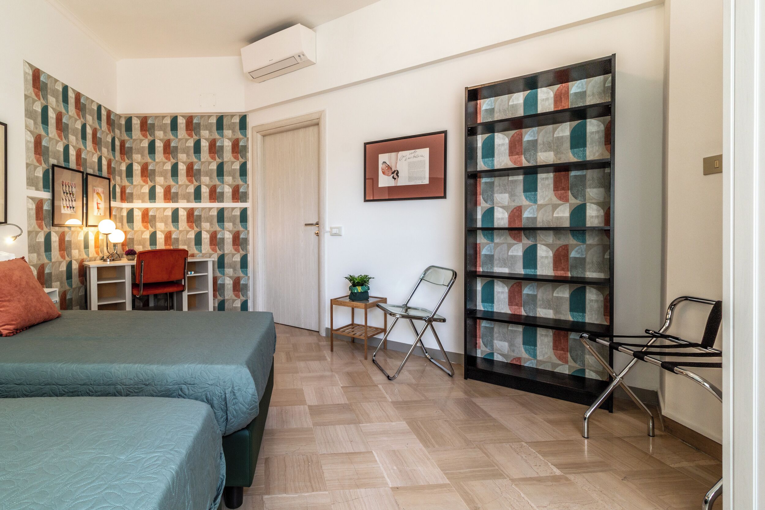 A compact room with a bed, a desk, and a chair. There is a bookshelf with colorful patterns and a framed picture on the wall.