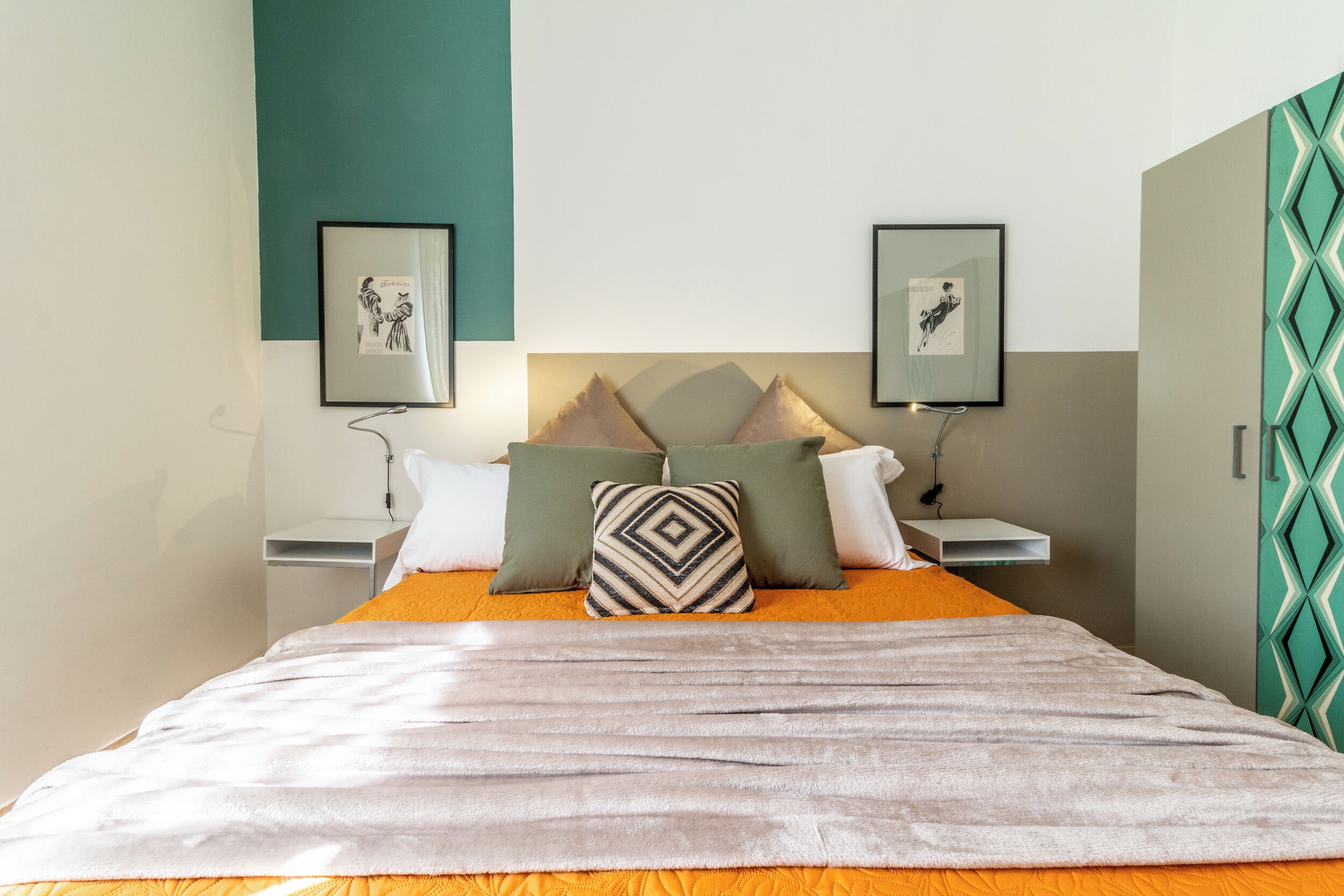A neatly made bed with a striped blanket, two pillows, and a headboard. There are two bedside tables with framed pictures and a wall-mounted shelf.