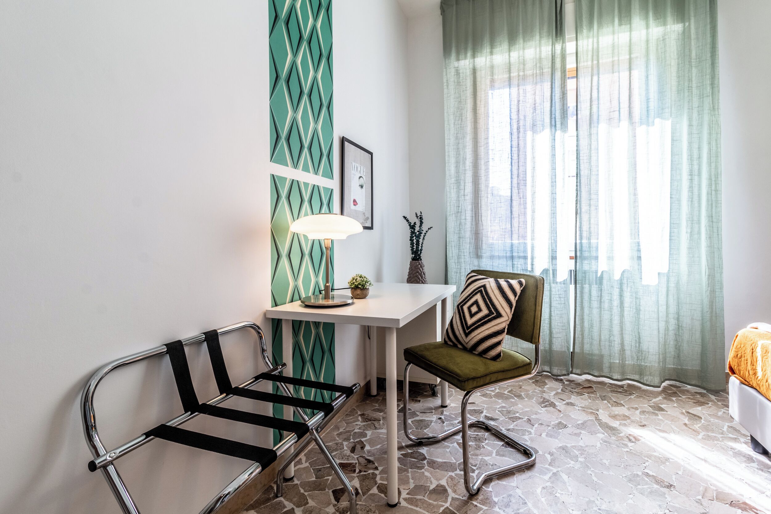 A room with a white desk, a green chair, a black chair, a lamp, and a bed with an orange blanket.