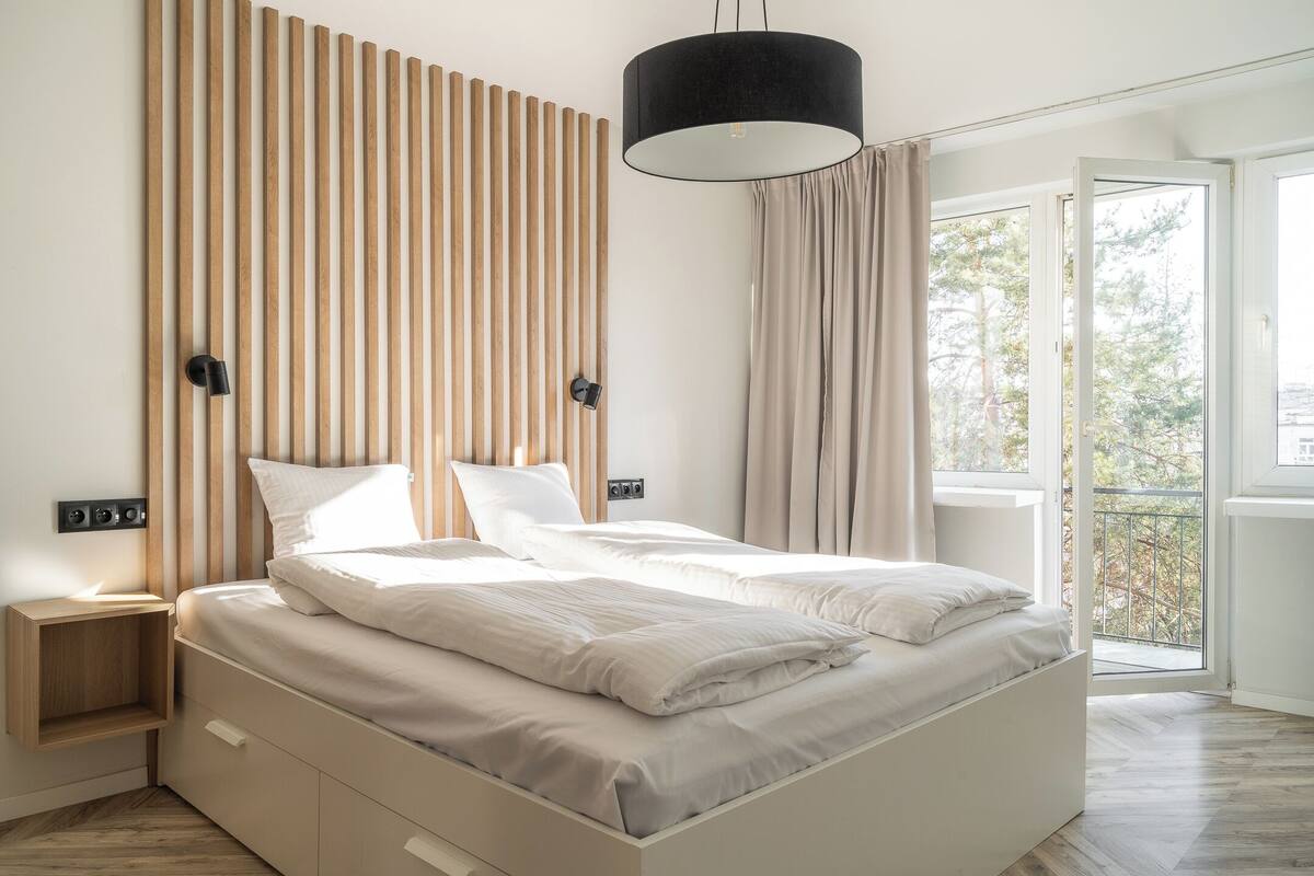 A modern bedroom with a large bed, wooden slats on the wall, a black pendant light, and a view of the outdoors through a large window.