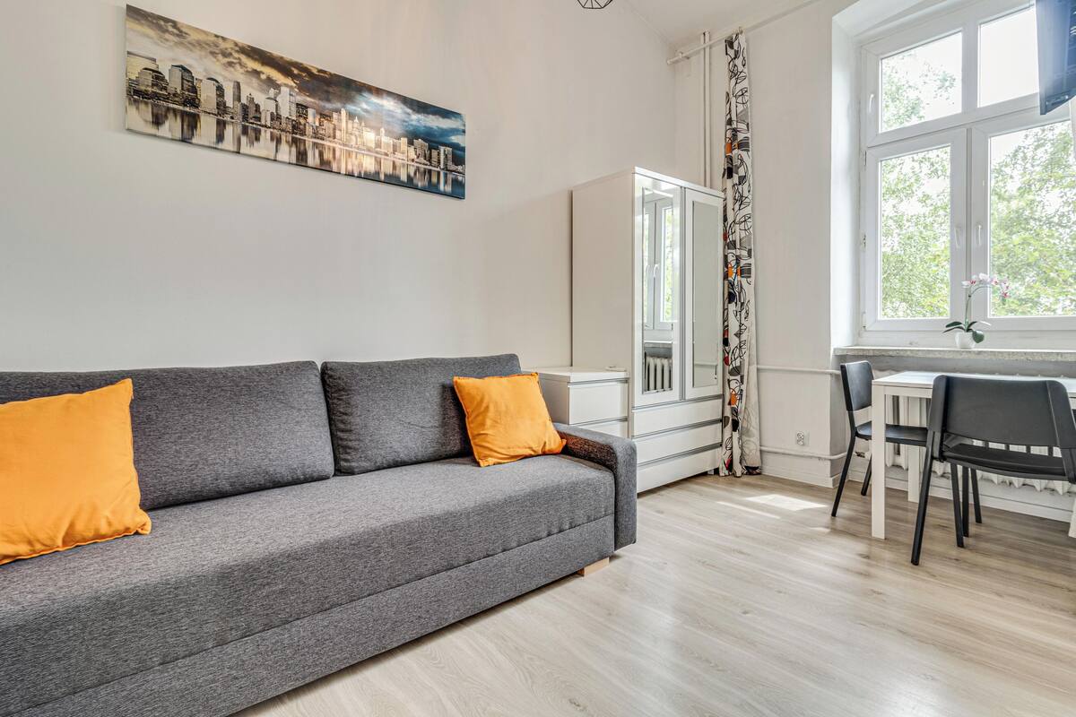 A modern living room with a grey sofa, orange cushions, a cityscape painting, a dining area with a white table and chairs, and a large window with a view of trees.