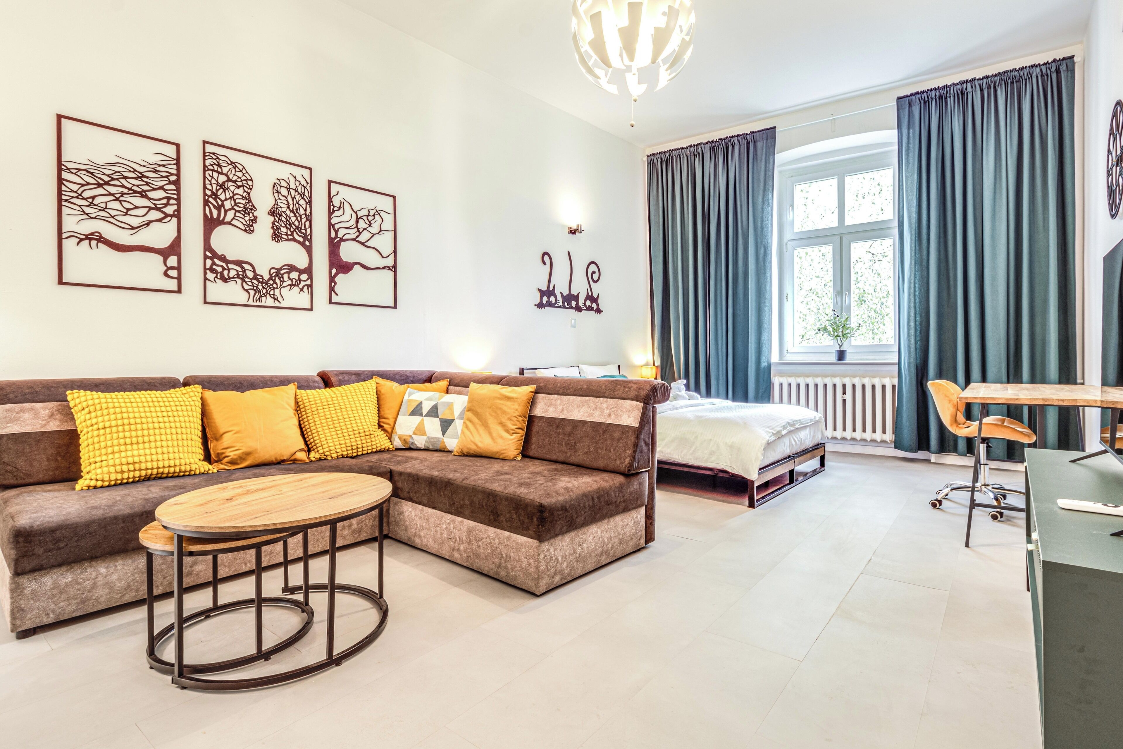 Ein modernes Wohnzimmer mit einem braunen Ecksofa, einem runden Couchtisch und einem Bett mit weißer Bettdecke. An der Wand hängen drei gerahmte Kunstwerke, ein Fenster mit Vorhängen und ein kleiner Tisch mit Stuhl.