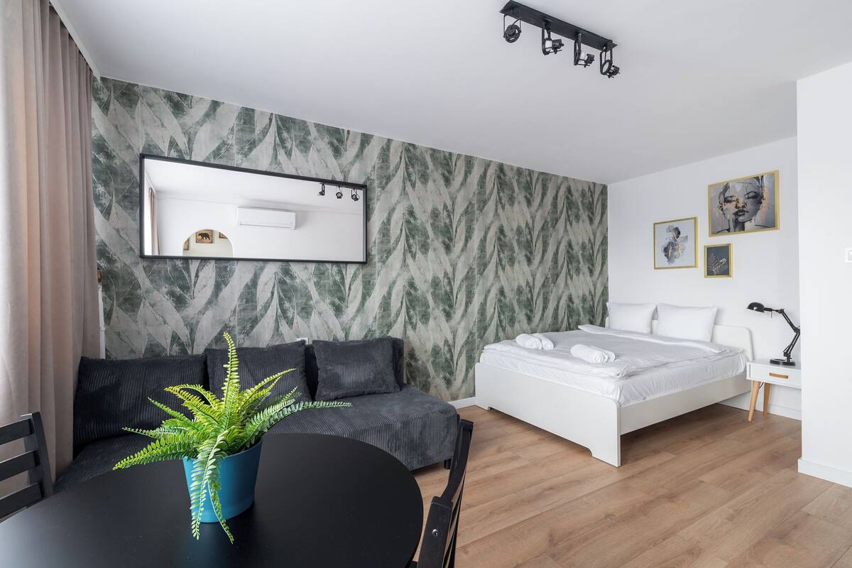 A modern hotel room with a bed, a sofa, a dining table with a plant, and a desk with a lamp.