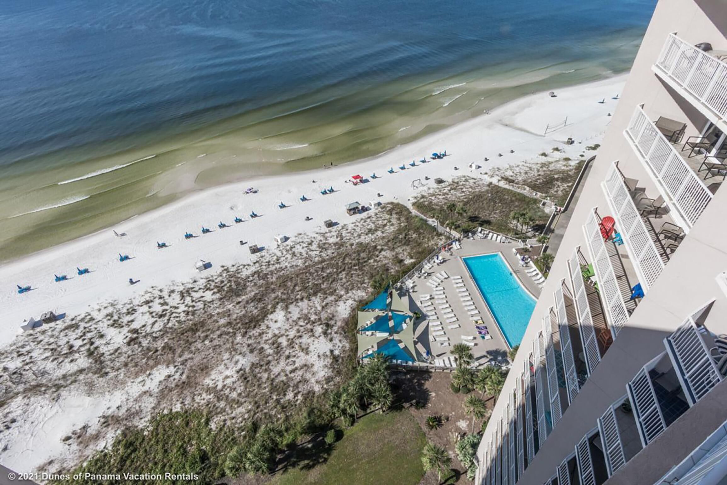 Vue aérienne d’un complexe hôtelier en bord de mer, avec une piscine, des parasols et un bâtiment doté de balcons.