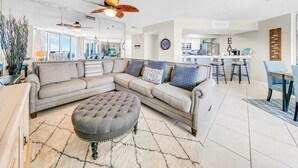 A modern living room with a sectional sofa, a round ottoman, and a ceiling fan. - Dunes of Panama 2104e (Panama City Beach)