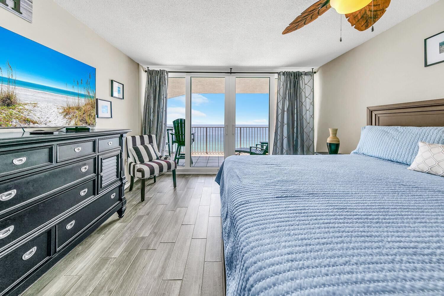 A bedroom with a bed, a dresser, a chair, a balcony with a view of the ocean, and a ceiling fan.
