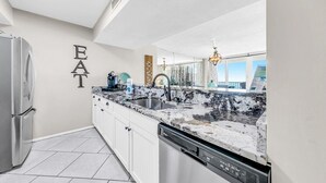 A modern kitchen with white cabinetry, a granite countertop, a stainless steel refrigerator, and a dishwasher. - Dunes of Panama 2104e (Panama City Beach)