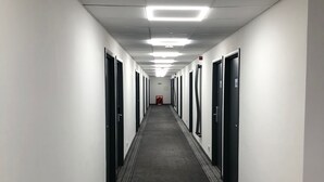 A long, narrow hallway with doors on both sides, carpeted floor, and fluorescent lighting. - My Motel (Paisley)