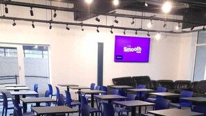 A spacious indoor area with multiple tables and chairs, a large screen displaying 'Smooth' on the wall, and a high ceiling with exposed beams and lighting fixtures. - My Motel (Paisley)