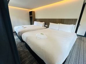 Habitación de hotel con dos camas, cada una con sábanas y toallas blancas, cabeceros de madera y suelo alfombrado.
