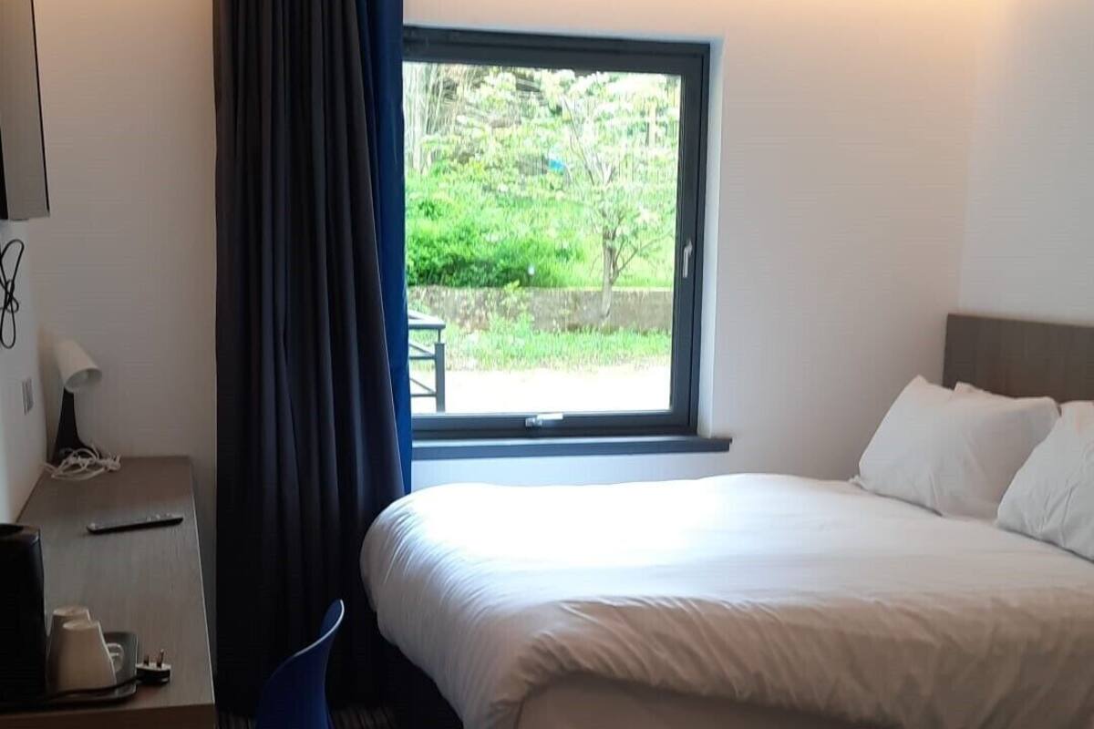 Une chambre d’hôtel comprenant un lit, un bureau, une chaise, une fenêtre donnant sur un espace verdoyant et un sol recouvert de moquette.