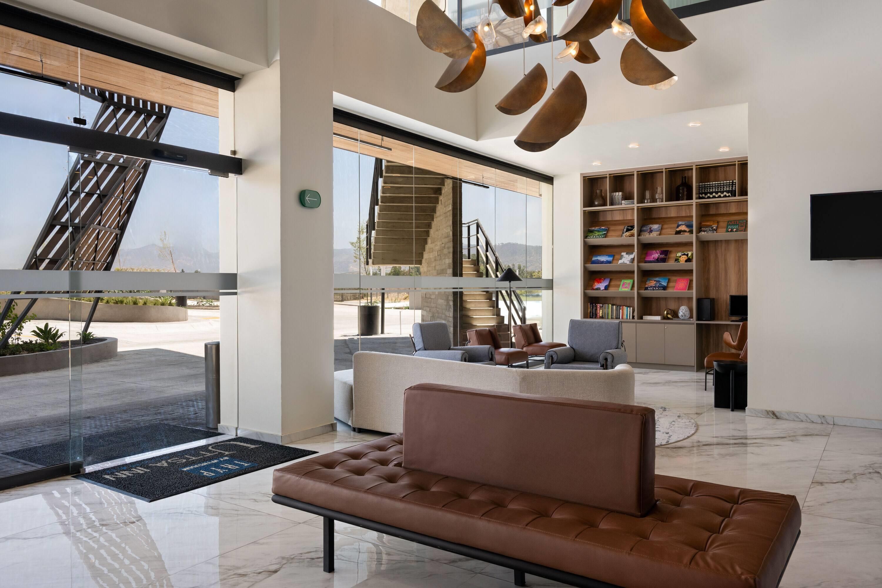 A modern interior with a leather sofa, a staircase, a bookshelf, and a large window offering a view of the outdoors.