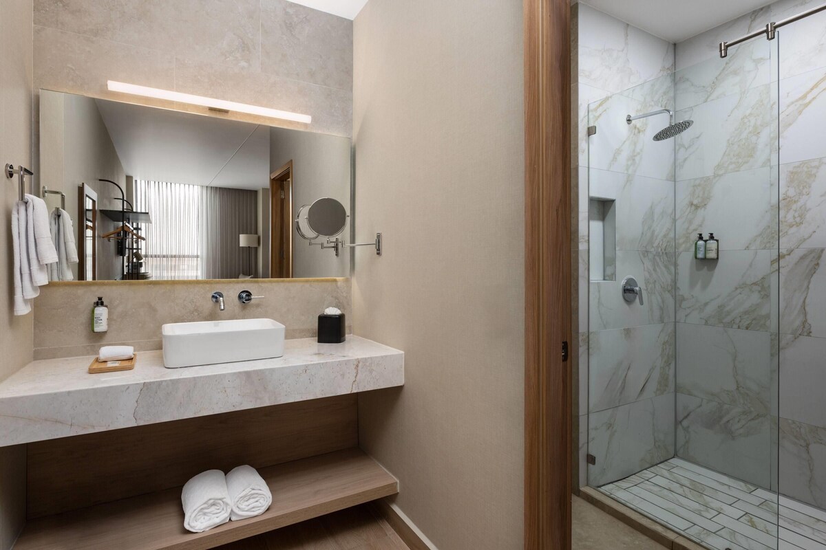 A modern bathroom with a marble sink, a large mirror, a handheld showerhead, and a glass-enclosed shower.
