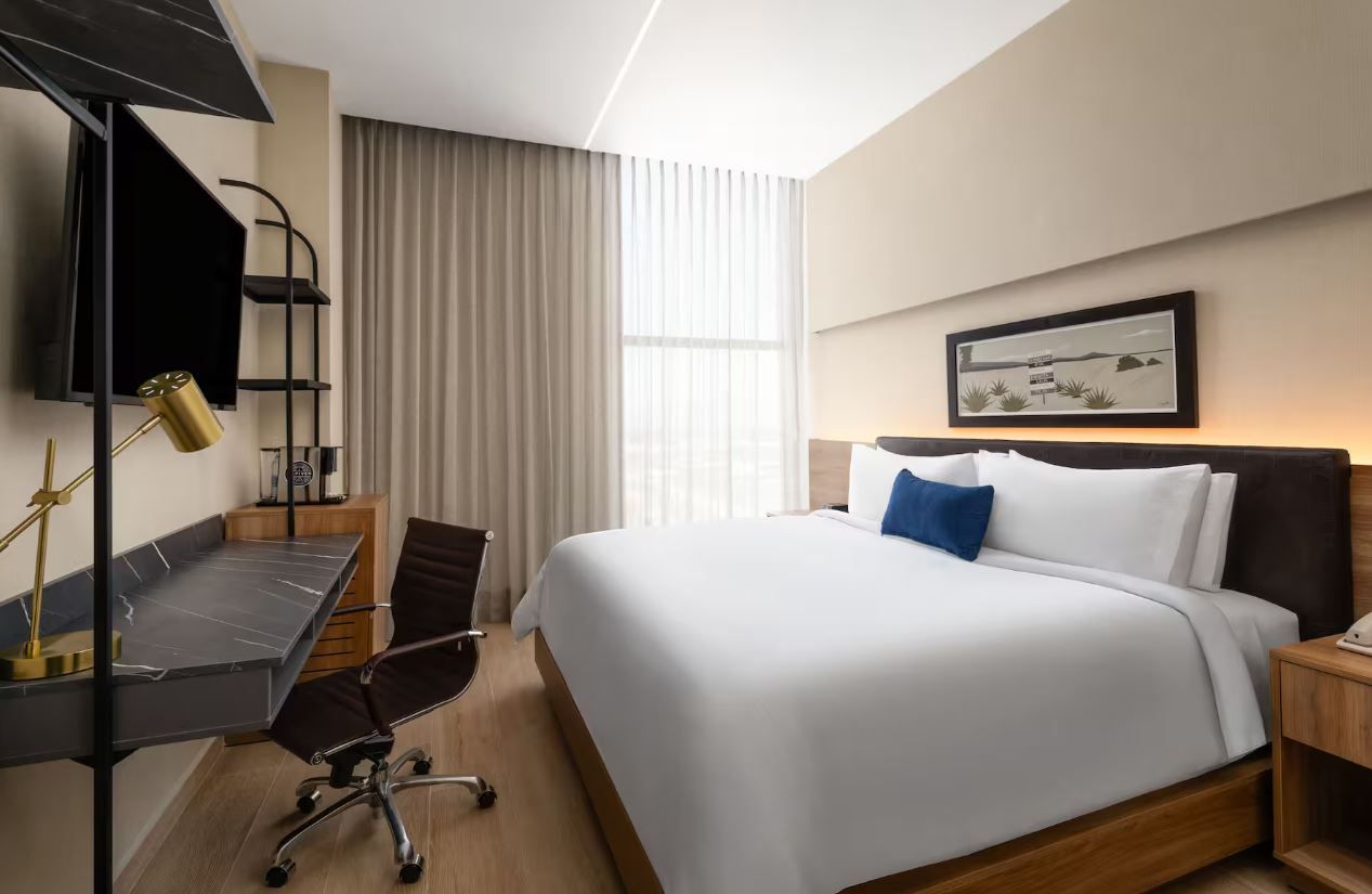 A modern hotel room with a large bed, a desk with a chair, a desk lamp, and a framed picture on the wall.
