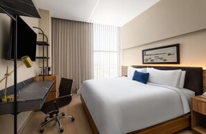 Ein modernes Hotelzimmer mit einem großen Bett, einem Schreibtisch mit Stuhl, einer Schreibtischlampe und einem gerahmten Bild an der Wand.
