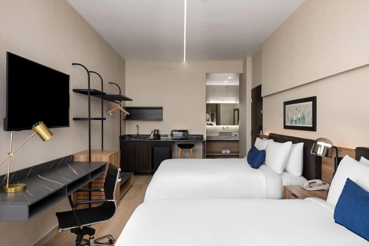 A hotel room with two beds, a desk with a computer, a TV, a kitchenette, and a bathroom mirror.